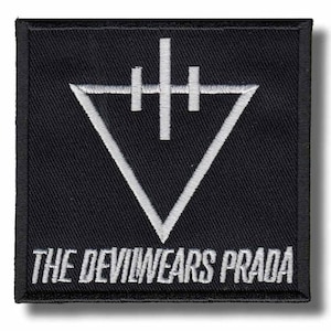 May include: Black square patch with white embroidered geometric design and text. The design features a downward-pointing triangle with a vertical line and two horizontal lines above it. The text reads "THE DEVIL WEARS PRADA".