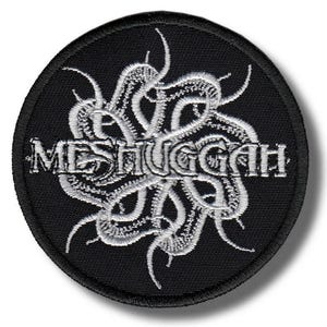 Op de afbeelding: Een ronde, zwarte patch met het woord "MESHUGGAH" in zilver geborduurd, omringd door een gestileerd, tentakelachtig ontwerp. De patch is waarschijnlijk voor kleding of accessoires.