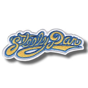 以下が含まれることがあります： 「Steely Dan」の文字がスタイリッシュな筆記体で刺繍されたパッチ。文字は金色の糸で、青い縁取りと白い縁があります。パッチは角が丸い長方形です。