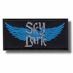 Puede incluir: Parche rectangular negro con el texto "Sky Lark" en una fuente estilizada, con alas azules. Las alas están detalladas con líneas de plumas individuales. El parche tiene un borde negro.
