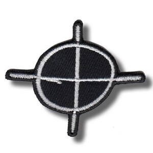 Puede incluir: Parche bordado en blanco y negro con un diseño de diana. El parche tiene un centro circular con una mira y cuatro brazos que se extienden. El diseño está cosido con hilo blanco sobre tela negra.