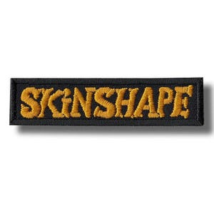 Op de afbeelding: Zwart rechthoekig embleem met het woord "SKINSHAPE" geborduurd in goudkleurig garen. De patch heeft een zwarte rand en is waarschijnlijk bedoeld om op kleding of accessoires te worden genaaid of gestreken.