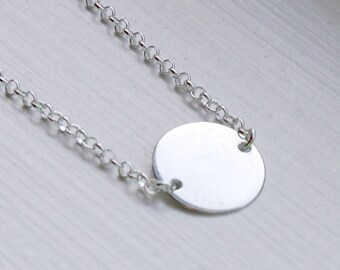 May include: A delicate silver necklace featuring a small, round pendant. The pendant is connected to a fine chain with a series of small, interconnected links. The necklace is displayed on a light-colored surface, highlighting its minimalist design.