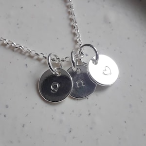Puede incluir: Collar de plata con tres dijes redondos. Los dijes están grabados con las letras 'g', 'n' y un corazón.