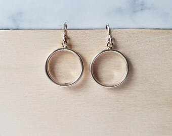 May include: A pair of silver hoop earrings. The earrings are round with a simple, minimalist design. They are hanging against a light wood surface with a blue and white background.