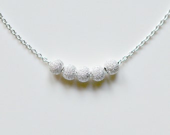 May include: A delicate silver necklace featuring five textured, spherical beads. The beads are evenly spaced along the chain, creating a subtle, elegant design. The necklace is displayed against a plain white background.