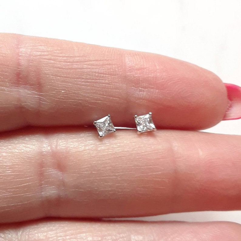 K&ouml;nnte beinhalten: Ein Paar kleiner, quadratisch geschliffener Ohrstecker mit klaren Edelsteinen. Die Ohrringe sind in einem silberfarbenen Metall gefasst und werden vor einem hellen Hintergrund pr&auml;sentiert.