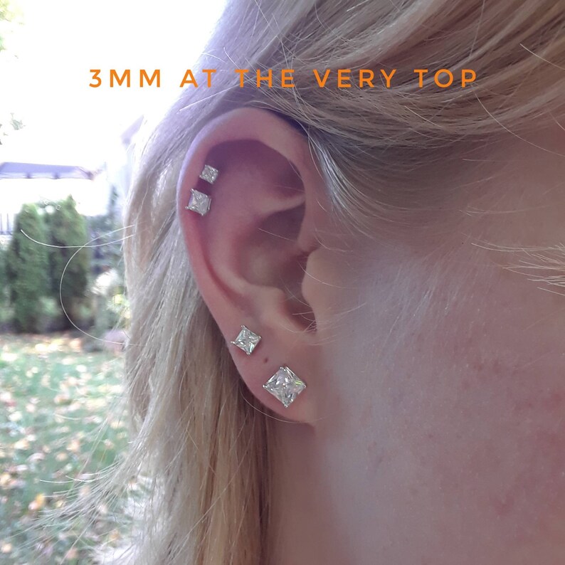 K&ouml;nnte beinhalten: Ein Ohr mit drei quadratischen, klaren Kristall-Ohrsteckern. Der Text oben lautet "3mm ganz oben". Die Ohrringe sind in einem silbernen oder wei&szlig;en Metall gefasst. Das Ohr ist vor einem verschwommenen Hintergrund im Freien.