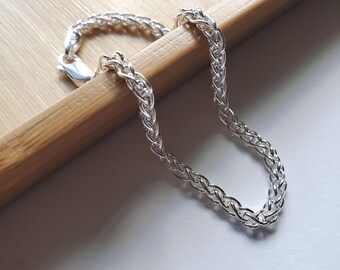 May include: A silver chain bracelet with a woven design. The bracelet is displayed on a light brown wooden surface, with a white background. The bracelet is made of a shiny, reflective metal and has a lobster clasp closure.