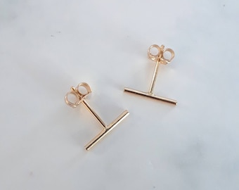 May include: A pair of gold-toned bar earrings with a simple, minimalist design. The earrings have a small post that goes through the earlobe and a short, straight bar that extends out from the post.