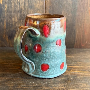 Puede incluir: Taza de cerámica hecha a mano con un diseño único. La taza presenta una base turquesa con lunares rojos y un borde marrón. El asa es de color turquesa a juego. Ideal para café, té o cualquier bebida.