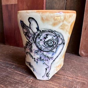 Puede incluir: Un vaso de cerámica con un diseño de conejo pintado a mano. El conejo está representado en tinta negra y morada, con un detalle circular en el ojo. El vaso tiene una base color crema con un borde marrón y una forma ligeramente trapezoidal.