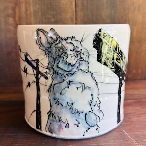 Puede incluir: Una taza de cerámica blanca con una ilustración en negro y azul de un conejo de pie sobre sus patas traseras. El conejo mira hacia la derecha y tiene una expresión de sorpresa. La taza también presenta una ilustración en negro y blanco de una línea eléctrica y un letrero amarillo con texto negro.