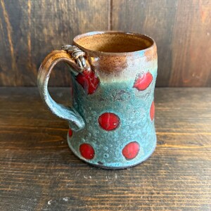 Pode incluir: Uma caneca de cerâmica artesanal com um design único. A caneca apresenta uma base azul-petróleo com grandes bolinhas vermelhas. A borda e a alça são marrons, com uma fixação de alça texturizada. Perfeita para café, chá ou qualquer bebida.