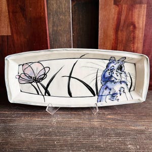 May include: Rectangular ceramic serving dish with a hand-painted design. The dish features a light blue and white color scheme, with a floral design on the left and a blue cat illustration on the right. The dish is supported by a clear stand.