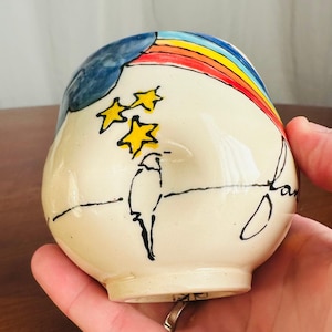 May include: A white ceramic mug with a hand-painted design of a rainbow, stars, and a bird. The mug has a blue cloud and the words "dream" written on it.