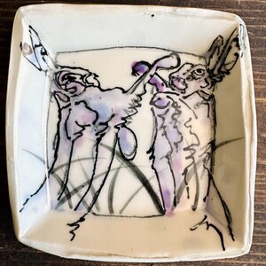 May include: Square ceramic dish with a hand-drawn illustration of two rabbits. The rabbits are depicted in black ink with purple and blue watercolor accents. The dish has a cream-colored base and a slightly raised edge.