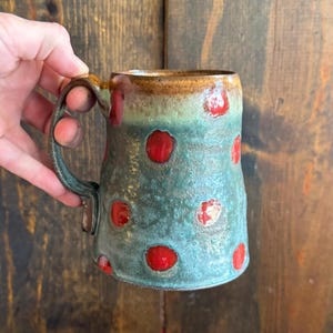Puede incluir: Taza de cerámica hecha a mano con un esmalte verde azulado y lunares rojos. La taza tiene un borde y un asa marrones, y una superficie texturizada única. Esta taza artesanal es perfecta para disfrutar de tus bebidas calientes favoritas.