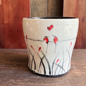 May include: A white ceramic cup with a black rim and base, decorated with red and black floral designs. The cup features a cracked glaze and a whimsical illustration of red hearts and flowers.