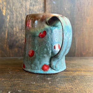 Puede incluir: Taza de cerámica hecha a mano con esmalte turquesa y lunares rojos. La taza tiene un borde y asa marrones, sobre un fondo de madera. Una pieza de cerámica única para disfrutar de bebidas.