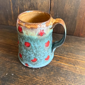 Puede incluir: Taza de cerámica hecha a mano con un esmalte turquesa y marrón. La taza presenta lunares rojos y un asa grande. El interior es marrón y el exterior tiene un acabado texturizado. Una pieza única para café o té.
