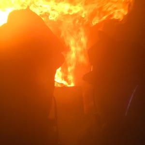May include: Two figures are silhouetted against a bright orange and yellow fire. The fire is intense and fills the background of the image.