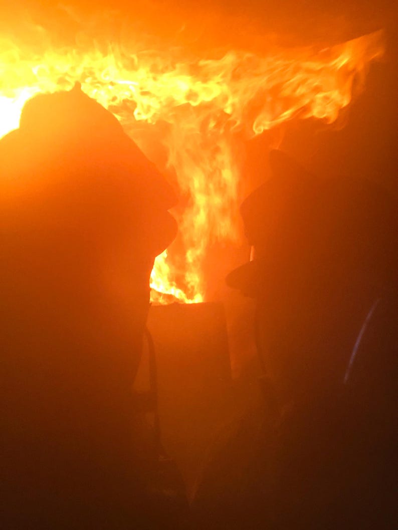 May include: Two figures are silhouetted against a bright orange and yellow fire. The fire is intense and fills the background of the image.