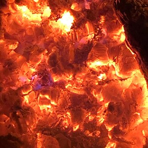 May include: Close-up of glowing red and orange embers in a fire pit. The embers are surrounded by dark grey ash.