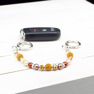 May include: A beaded keychain with a variety of beads in shades of gold, brown, and pearl. The keychain is attached to a car key and has two silver-colored clasps.