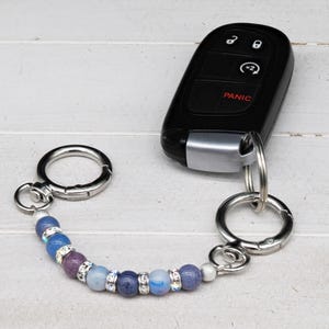 May include: A beaded keychain with a black car key fob. The keychain features a series of blue, purple, and clear beads with silver accents. The car key fob is black with silver trim and has buttons for locking, unlocking, and a panic button.
