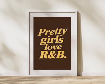 May include: A framed art print with the text "Pretty girls love R&B" in a yellow, retro font on a dark brown background. The print is in a white frame and leans against a white wall.