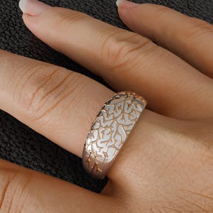Könnte beinhalten: Ein roségoldener Ring mit einem weißen, filigranen, durchbrochenen Design. Das Band des Rings weist ein detailliertes Muster aus miteinander verbundenen Formen auf, das ein zartes und ornamentales Aussehen erzeugt. Der Ring wird am Finger getragen.