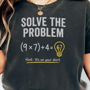 Pode incluir: Camiseta cinza escuro com o texto "SOLVE THE PROBLEM" acima de uma equação matemática e um gráfico de lâmpada. A equação é (9x7)+4=67. Abaixo, o texto "Hint: It's on your shirt."