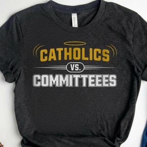 Op de afbeelding: Donkergrijs t-shirt met de tekst "Catholics vs. Committeees" in geel en wit. Het shirt is gestyled met een blauwe jeans en witte sneakers. Het shirt heeft een ronde hals en korte mouwen.