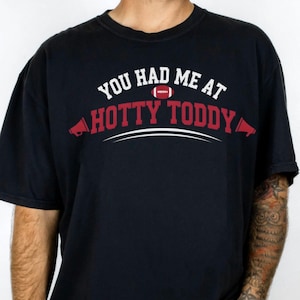 Puede incluir: Camiseta negra con el texto "YOU HAD ME AT HOTTY TODDY" en blanco y rojo. El diseño incluye un gráfico de fútbol americano e ilustraciones de megáfonos. La camiseta es de estilo casual.