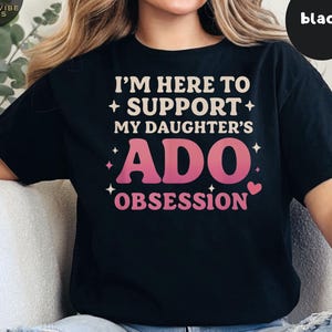Puede incluir: Camiseta negra con el texto "I'M HERE TO + SUPPORT + MY DAUGHTER'S ADO OBSESSION" en blanco y rosa. La camiseta tiene cuello redondo y mangas cortas. Un círculo negro con la palabra "black" está en la esquina superior derecha.