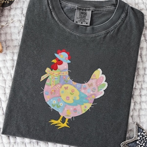 Puede incluir: Camiseta gris oscuro con un diseño de gallina de retazos coloridos. La gallina lleva un sombrero azul y un lazo amarillo. La palabra "pepper" está en un cuadro gris en la esquina superior derecha.