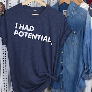 Könnte beinhalten: Ein marineblaues T-Shirt mit dem weißen Text "I HAD POTENTIAL". Das Shirt hängt an einem Holzbügel mit anderen Kleidungsstücken im Hintergrund. Das Shirt ist unten geknotet.