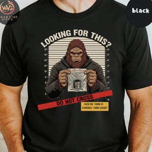Könnte beinhalten: Schwarzes T-Shirt mit einem Bigfoot-Motiv, das einen Beutel mit einer 10-mm-Stecknuss hält. Der Text "LOOKING FOR THIS?" steht über der Grafik. Ein rotes "DO NOT CROSS"-Band und eine Akte sind darunter.
