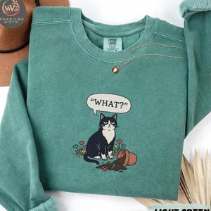 May include: Light green sweatshirt featuring a black and white cat illustration with the word "WHAT?" in a speech bubble. The cat sits near a tipped-over flower pot. A gold necklace is visible.