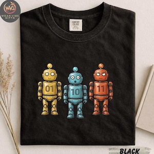 Könnte beinhalten: Schwarzes T-Shirt mit drei Cartoon-Robotern in Gelb, Blau und Orange. Jeder Roboter hat eine andere Nummer auf der Brust: 01, 10 und 11. Das Shirt hat ein Comfort Colors-Etikett und das Wort "BLACK" ist unten aufgedruckt.