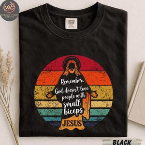 May include: Black t-shirt with a graphic of Jesus in a colorful sunset circle. The text reads "Remember, God doesn't love people with small biceps. JESUS." The shirt has a Comfort Colors tag.