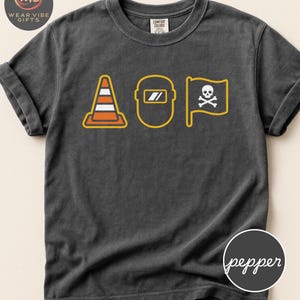May include: Dark gray t-shirt with three yellow and orange graphics: a traffic cone, a welding mask, and a pirate flag with a skull and crossbones. The shirt has rolled-up sleeves and a Comfort Colors tag.