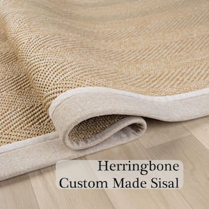 May include: Close-up of a rolled-up sisal rug with a herringbone pattern. The rug has a natural beige color with a lighter beige border. The text "Herringbone Custom Made Sisal" is visible.
