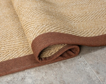 May include: A rolled-up, rectangular rug with a natural, woven texture. The rug features a light tan center and a dark brown border. The rug is made of a durable material and is suitable for indoor use.