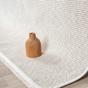 a wooden vase placed on a beige woven rug with a white fringe.