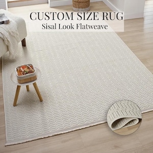 Puede incluir: Una alfombra rectangular de color blanquecino con un patrón geométrico. Se lee "CUSTOM SIZE RUG" y "Sisal Look Flatweave". Un primer plano muestra la textura de la alfombra. La alfombra está sobre un suelo de madera clara.