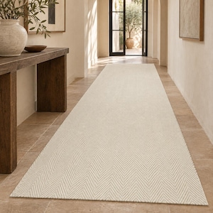 Puede incluir: Una alfombra de pasillo larga y rectangular con un patrón de espiga en color beige claro y crema. La alfombra está colocada sobre un suelo de baldosas, extendiéndose desde el primer plano hacia una puerta al fondo, mostrando una textura tejida.