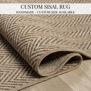 Op de afbeelding: Een opgerolde, op maat gemaakte sisal vloerkleed met een visgraatpatroon. Het vloerkleed heeft een natuurlijke beige kleur met donkerbruine accenten. De tekst "CUSTOM SISAL RUG" en "HANDMADE - CUSTOM SIZE AVAILABLE" staat bovenaan de afbeelding.