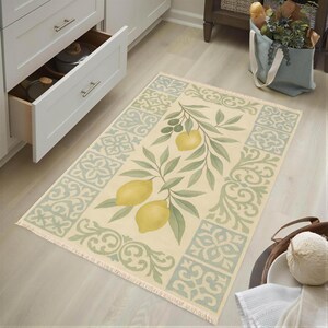 a kitchen with a rug on the floor, which has a lemon tree design. The rug is placed near a white cabinet with a drawer. There is also a bag of fruit and a bowl of fruit on the floor.